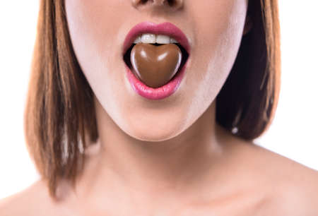 Sweet temptation. Beautiful woman holding heart-shaped chocolate candy in her mouth.の写真素材