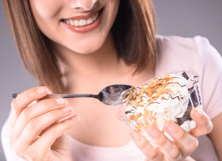 Sweet temptation. Young smiling woman tasting creamy dessert over gray background. Close-up.