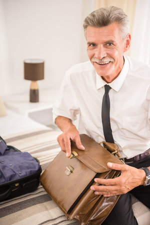 Mature man packing his things into the briefcase while sitting on bed at the hotel room.の写真素材