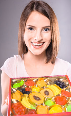 Sweet-tooth. Beautiful woman holding box of  fruit jellies over gray background.の写真素材