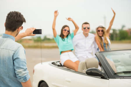 Happy friends sitting in cabriolet. Young man taking photo at his phone.の写真素材