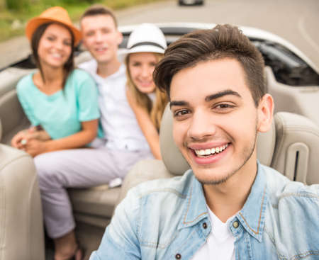Handsome man sitting in cabriolet, looking at camera and smiling against his friends on rear seats.の写真素材