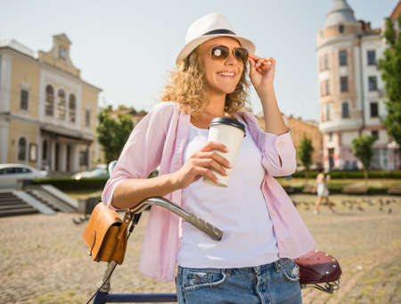 Young woman in sunglasses with bicycle holding cup of coffee and looking away.の写真素材