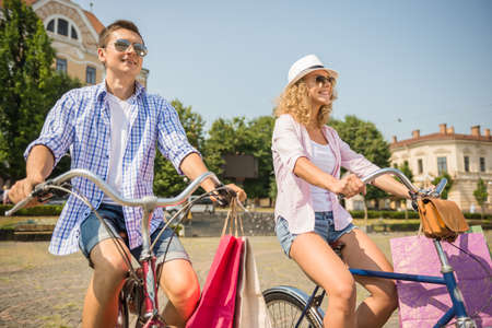 Happy couple in sunglasses with shopping bags riding on bicycles on the city street.の写真素材
