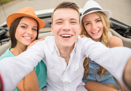 Three cheerful friends taking selfie while sitting in the cabriolet on rear seats. Front view.の写真素材