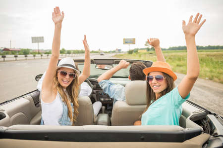 Shot back. Group of friends traveling on the cabriolet with hands up. Girls looking at camera.の写真素材