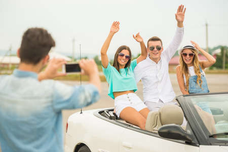 Happy friends sitting in cabriolet. Young man taking photo at his phone.の写真素材