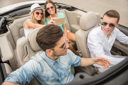 Group of happy friends sitting in the cabriolet. Beautiful girls taking a selfi on rear seats.の写真素材