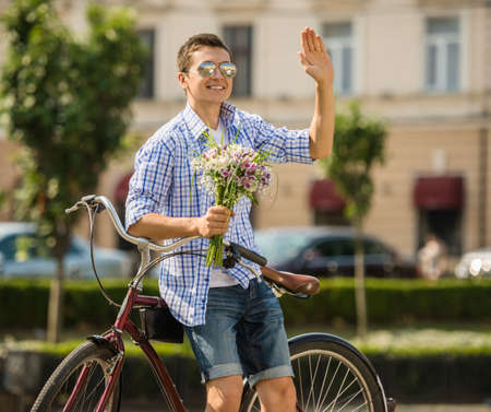 Young man in sunglasses with bicycle holding flowers and waiting his girlfriend at the city street.の写真素材