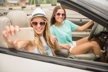 Two attractive girls taking selfie while driving through countryside in cabriolet.の写真素材
