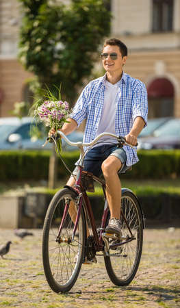 Young man in sunglasses with flowers riding on bicycle at the city street in sunny day.の写真素材
