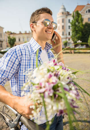 Portrait of cute man in sunglasses with bicycle holding flowers and talking on phone at the city.の写真素材