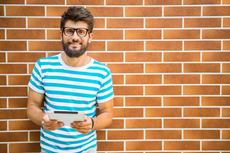 Smiling young man is standing near the wall and holding a digital tablet.の写真素材