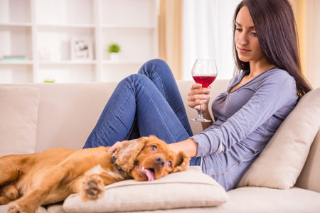 Young woman is relaxing at home with a glass of wine, and her dog is lying near on sofa.の写真素材
