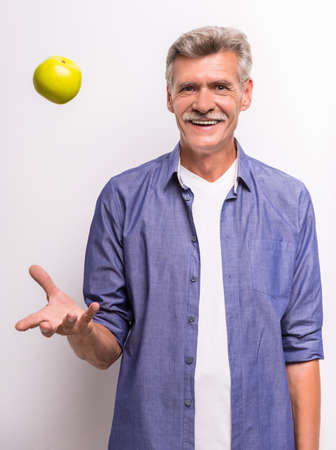 Smiling senior man in casual throwing a green apple up and smiling while standing against grey background.の写真素材