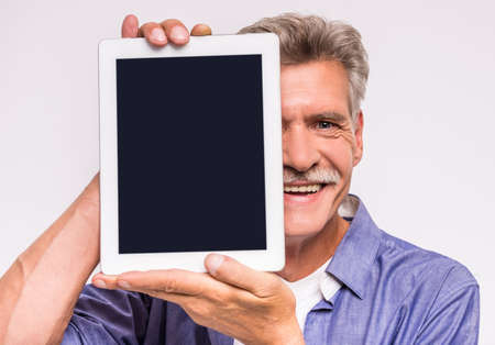 Senior mature man is showing digital tablet and smiling while standing against grey background.の写真素材