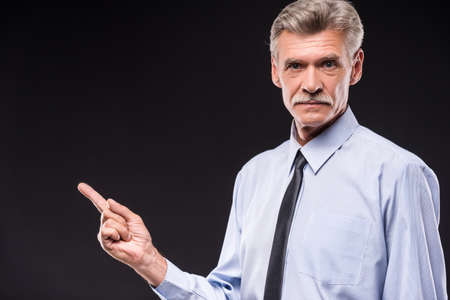 Senior man in shirt is pointing with finger away, dark background.の写真素材