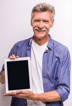 Senior mature man is showing digital tablet and smiling while standing against grey background.の写真素材