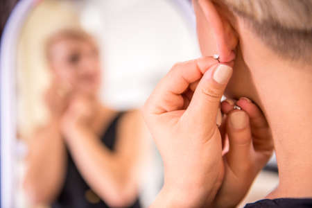 Beautiful young woman putting on her a new earrings and smiling while looking at the mirror.の写真素材