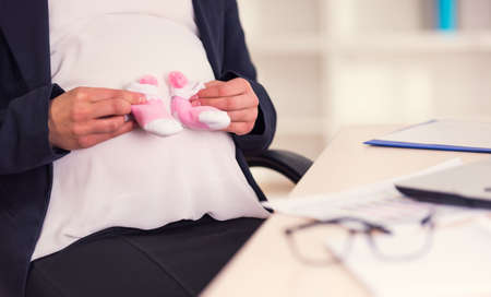 Happy pregnant woman looks at socks for future baby while working in the officeの写真素材