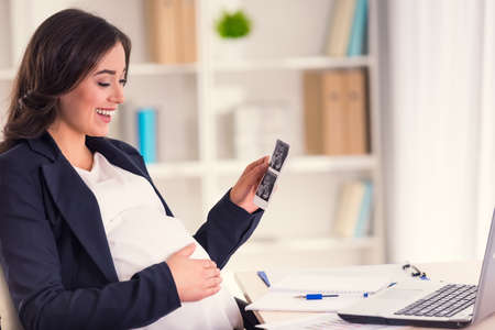 Happy pregnant woman watching ultrasound while working in the officeの写真素材