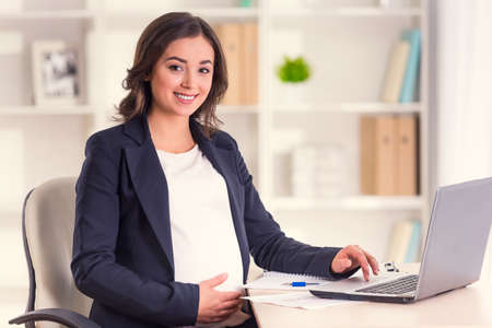 Happy pregnant woman working on a laptop in the office holding bellyの写真素材
