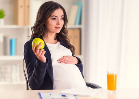 A young pregnant woman happy eating an apple and drinking juice sitting in the officeの写真素材
