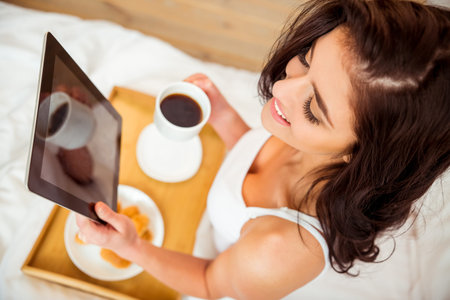 Beautiful girl in a white shirt using a tablet during breakfast in bedの写真素材