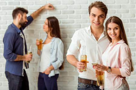Smiling young couple holding a bottle of beer and a smartphone, the other a young couple with a...