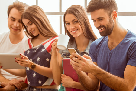 Four young attractive people, keeping at hand tablet computer, cup, smartphone and notebook, sit on...