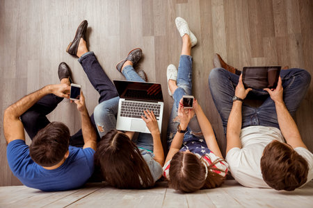 Group of attractive young people sitting on the floor using a laptop, Tablet PC, smart phones,...