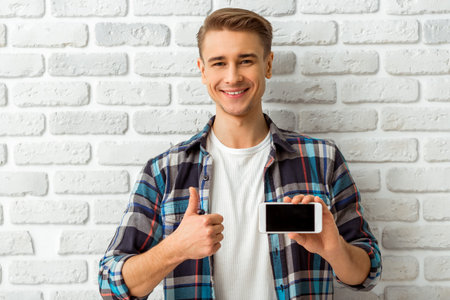 Beautiful young man in casual wear holding the phone in hand  on the background white brick wall