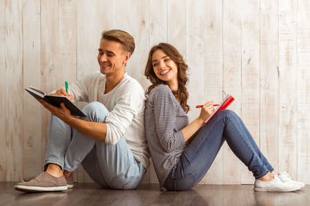 Beautiful young couple writing in a notebook sitting on wooden floor at homeの写真素材