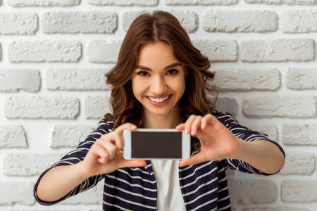 Beautiful young woman making self-portrait on a smartphone on a brick wall background