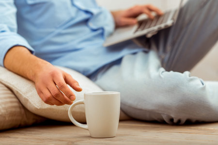 Young handsome man in the blue shirt and jeans using a laptop, next to a cup sitting on the floor on...