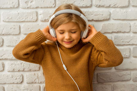 Portrait of pretty little blonde girl listening to music and smilng while sitting against white brick wallの写真素材