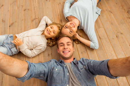 Top view of beautiful young family looking in camera and smiling while lying on wooden floor at home. Father making selfie.の写真素材