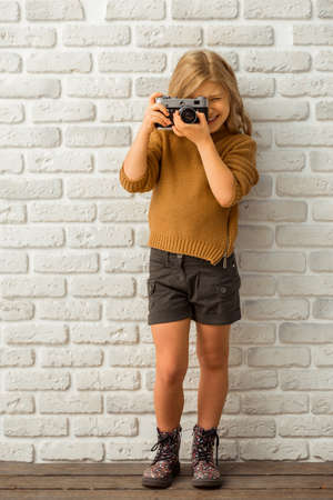 Pretty little blonde girl smiling and making a photo while standing against white brick wallの写真素材