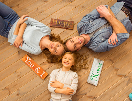 Top view of beautiful young family looking in camera and smiling while lying on wooden floor at...