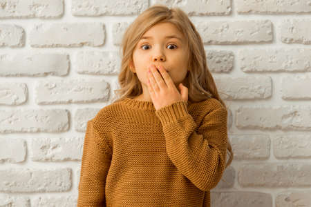 Portrait of a pretty little blonde girl showing emotions, covering her mouth and looking in camera while standing against white brick wallの写真素材