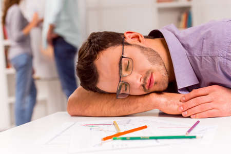 Young attractive businessman in casual clothes and eyeglasses sleeping in office, in the background man and woman talkingの写真素材