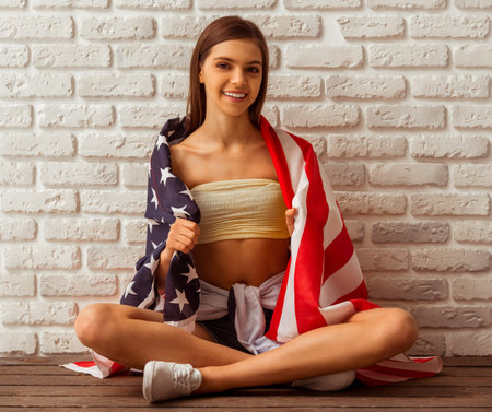 Cute teenage girl in a top and shorts sitting covered with an American flag against white brick wall, looking in camera and smilingの写真素材