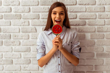 Cute teenage girl holding a lollipop, looking in camera and smiling, standing against white brick wallの写真素材