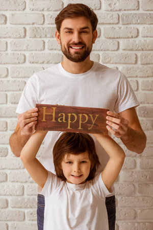 Portrait of a handsome young father and his cute little son holding a wooden plate "happy", looking in camera and smiling. Both in white t-shirts and jeans, standing against white brick wall.の写真素材