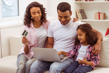 Cute little Afro-American girl and her beautiful young parents using a laptop and doing shopping online while sitting on a sofa at home. Mother holding a credit card.の写真素材
