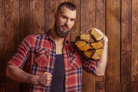 Stylish man with beard in casual shirt holding firewood and looking at camera, standing on a wooden...