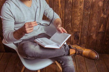 Stylish man in gray sweater holding eyeglasses and reading a book, sitting on a chair against wooden...