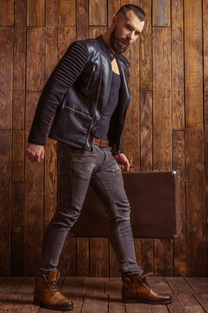 Stylish man with beard in leather jacket holding a suitcase and looking at camera, standing on a wooden backgroundの写真素材