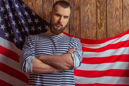 Stylish man with beard in striped vest looking at camera, standing on a wooden background with American flagの写真素材