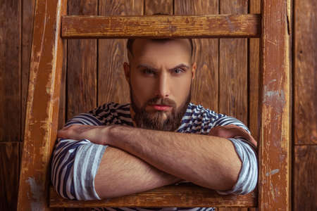 Stylish man with beard in striped vest leaning on a ladder, looking at camera, standing on a wooden background, close-upの写真素材